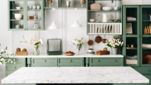 Clean and empty marble countertop, green vintage kitchen furniture with lots of flowers and bowl of strawberries, pair of white hanging pendant lights, various crockery in blurred background