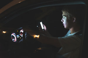 The images displays a distracted teenager driving a car with his cell phone in his hand. The light from the screen of the phone is illuminating his face.
