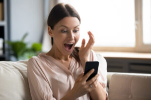 Amazed young woman sit on couch at home feel shocked reading unexpected good news on cellphone gadget, stunned millennial girl surprised by online lottery win on smartphone, luck concept