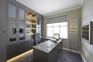a beautiful office in a luxury house decorated in tones of grey with generous use of grey mock-snakeskin leather (backdrop to the display shelves, the lower cupboard doors, the desk sides and surround to the wall-mounted TV). The display cabinet is lit with LED lights together with the base of the cupboards. The surface of the desk is a large piece of glass.